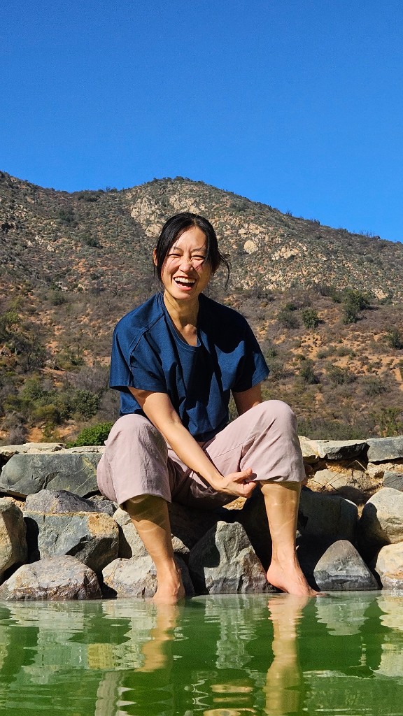 Serok Park sonriendo junto al agua, con los pies en el río y las montañas al fondo